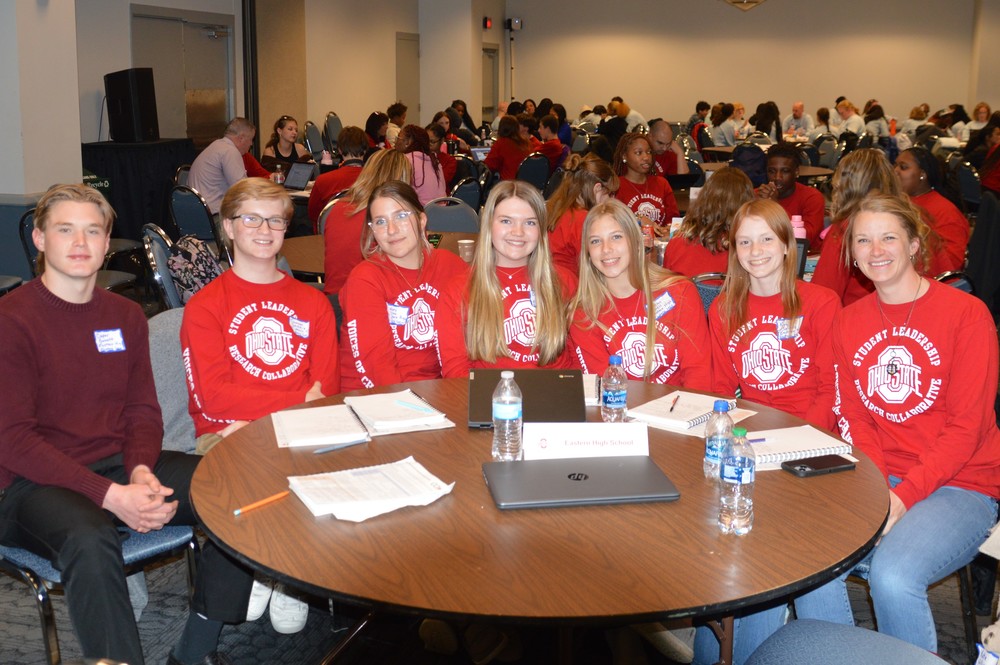 Six Eastern students and their teacher wear red long sleeve shirts and smile at a table during a conference in Cleveland.