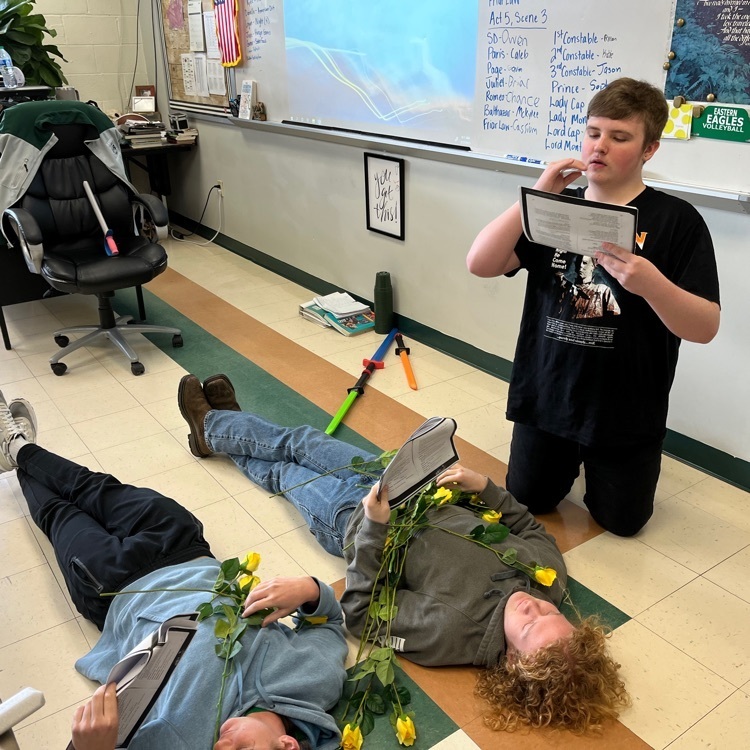 student drinks pretend potion above two students on floor with flowers scattered around them.