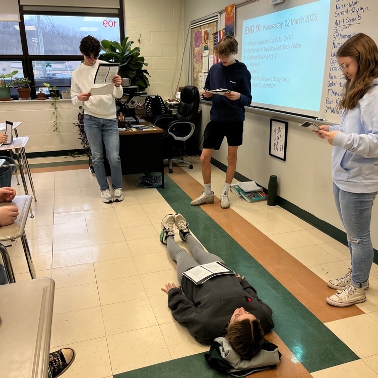 three students stand to read Romeo and Juliet while one lies on the floor.